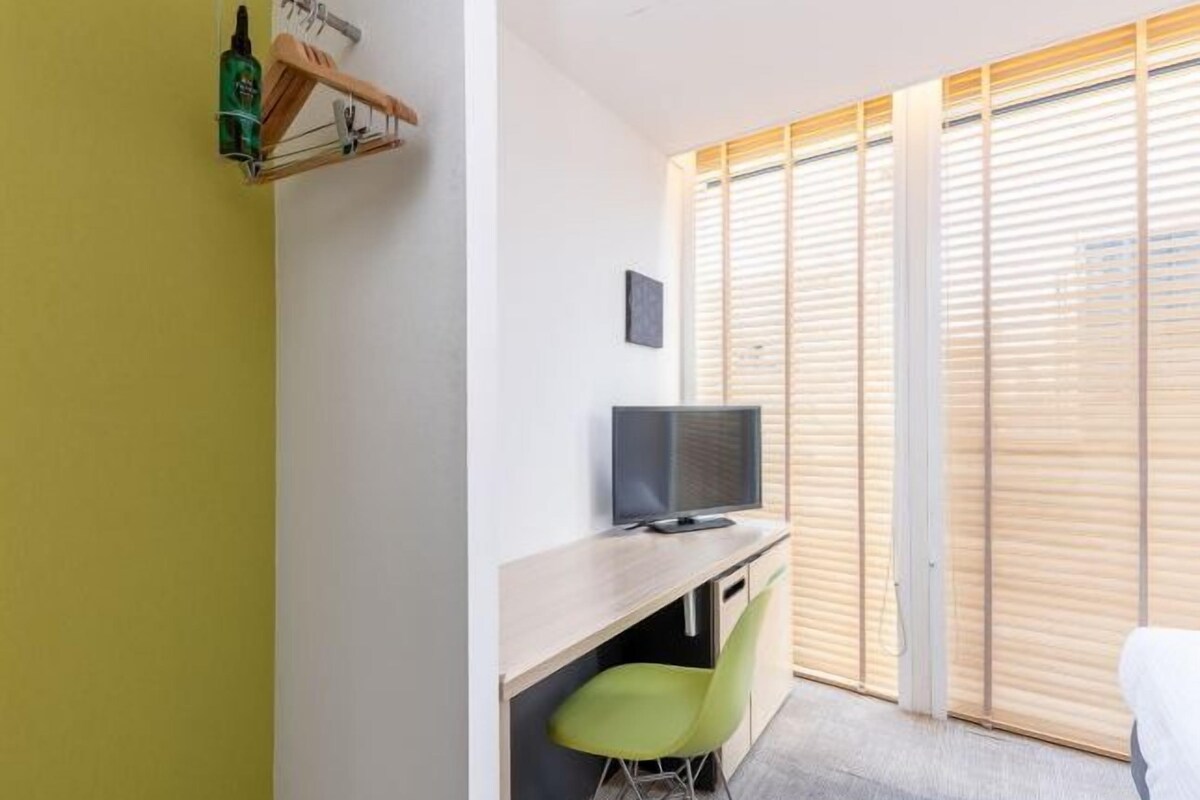 A room with a wooden desk, a green chair, a television, and a window with blinds.