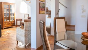 A spacious living area with a high ceiling, a wooden cabinet with glass doors, wicker chairs, a glass table, and a staircase with a railing.