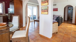 A spacious living area with a glass dining table, wicker chairs, a sofa, and a kitchen area with cabinets and a refrigerator.