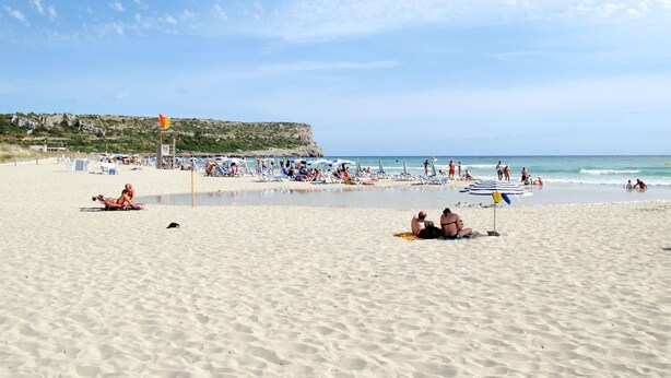 A beach with people sunbathing and swimming, a lifeguard tower, and a rocky cliff in the background.