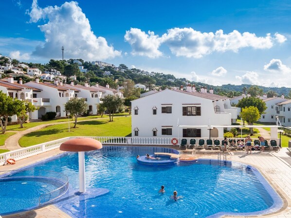 A swimming pool with a circular hot tub, surrounded by a white building with a tiled roof, set against a backdrop of a hilly landscape.