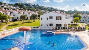 A swimming pool with a circular hot tub, surrounded by a white building with a tiled roof, set against a backdrop of a hilly landscape.