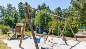 A playground with swings and a slide, surrounded by trees and a small building.
