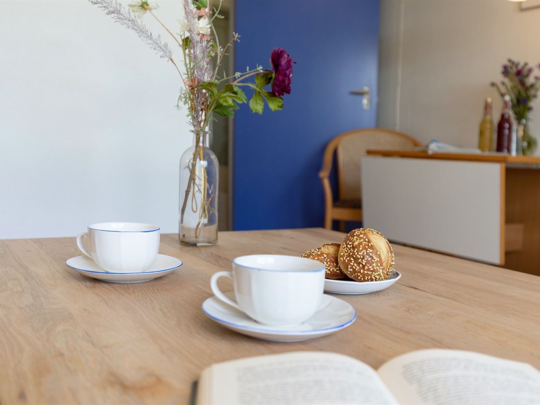 A wooden table with two white cups on saucers, a plate with sesame seed buns, a vase with flowers, and an open book.