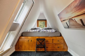 A loft bedroom with a sloped ceiling, a wooden dresser, a small stool, a bed with a gray and purple blanket, and a large window.