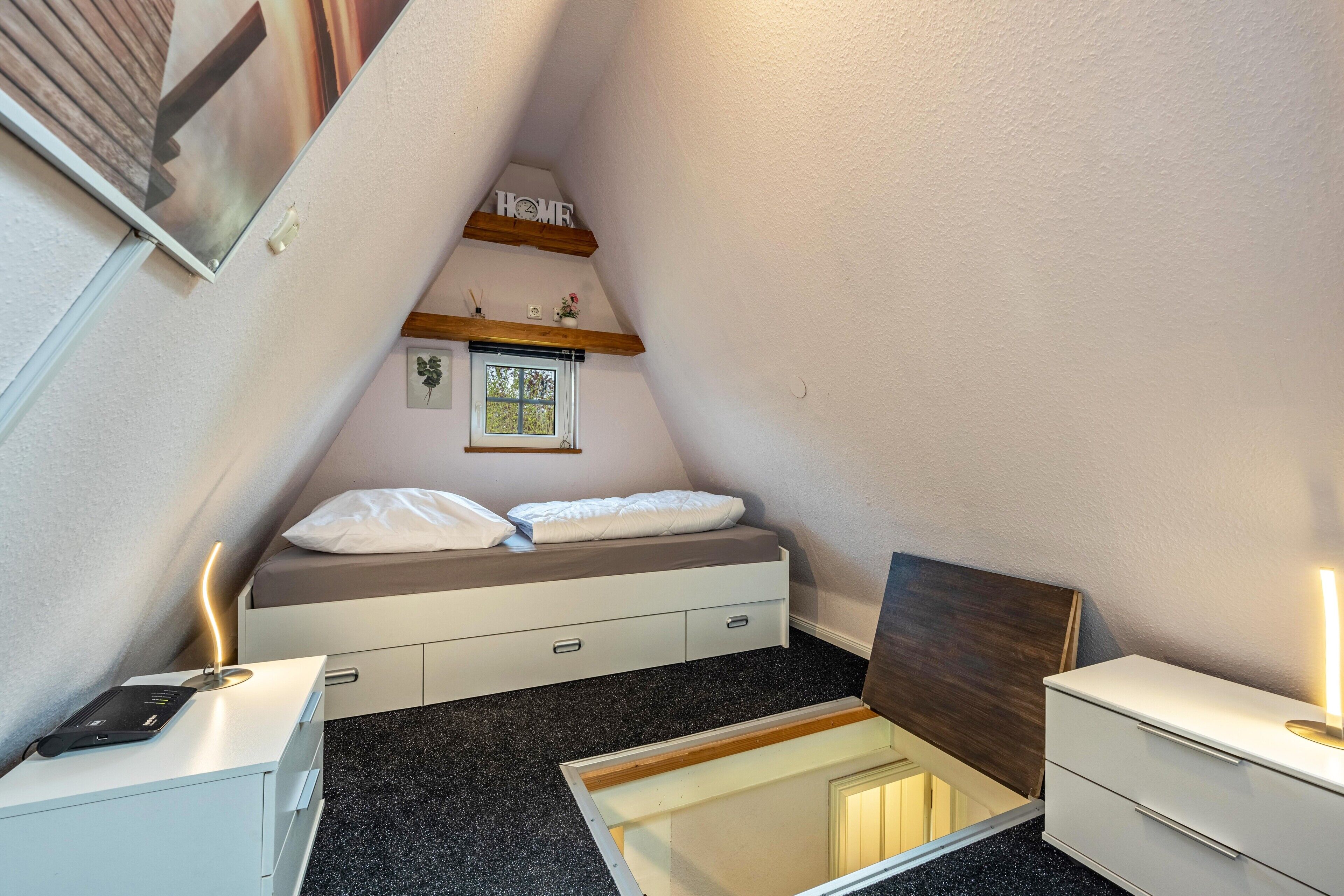 A bedroom with a loft bed, a bedside table, a nightstand, a small window, and a wall-mounted shelf with a 'HOME' sign.