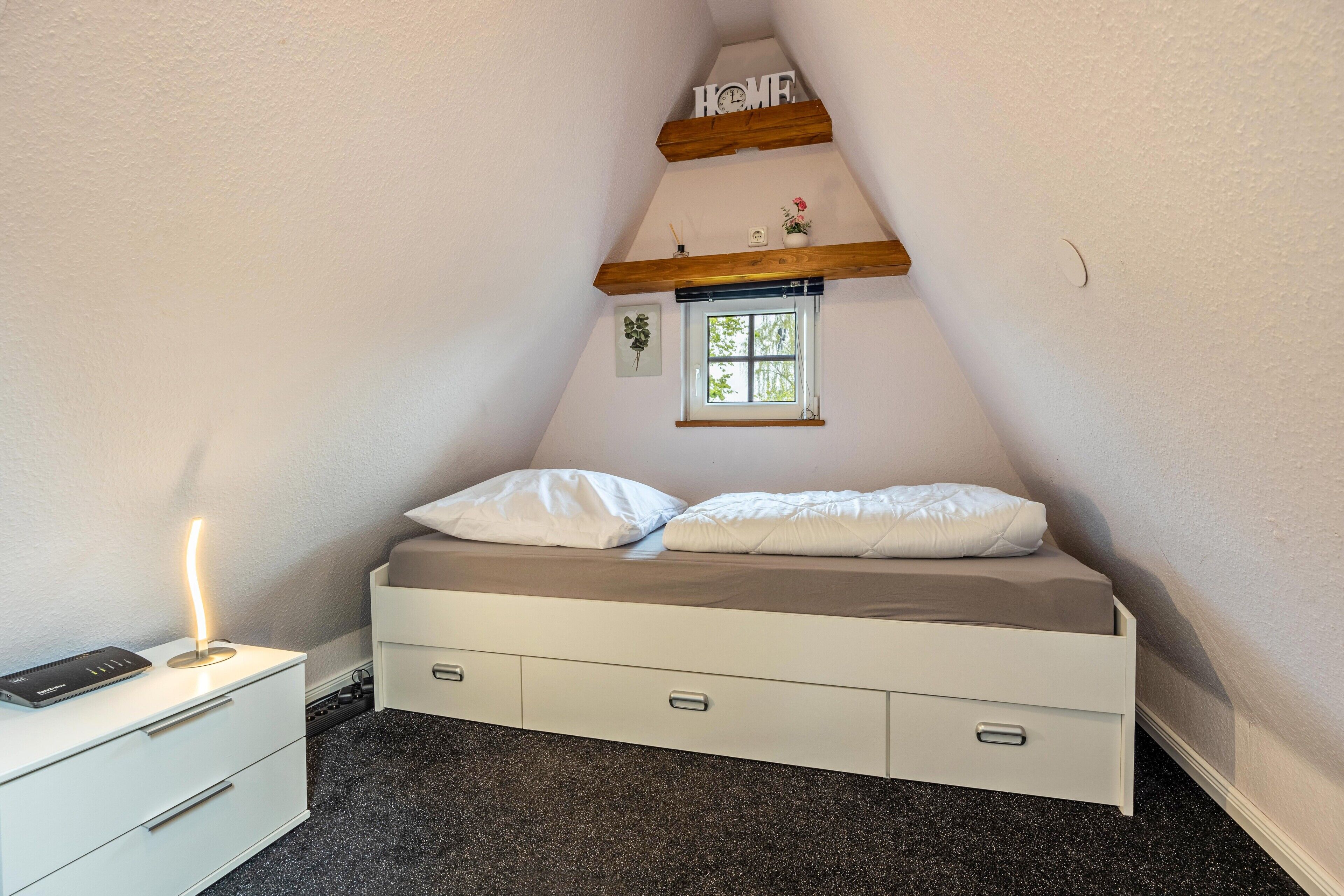 Ein Schlafzimmer mit schräg versiegelter Decke, einem Bett mit weißer Bettwäsche, einem Nachttisch mit Lampe und einem kleinen Fenster.