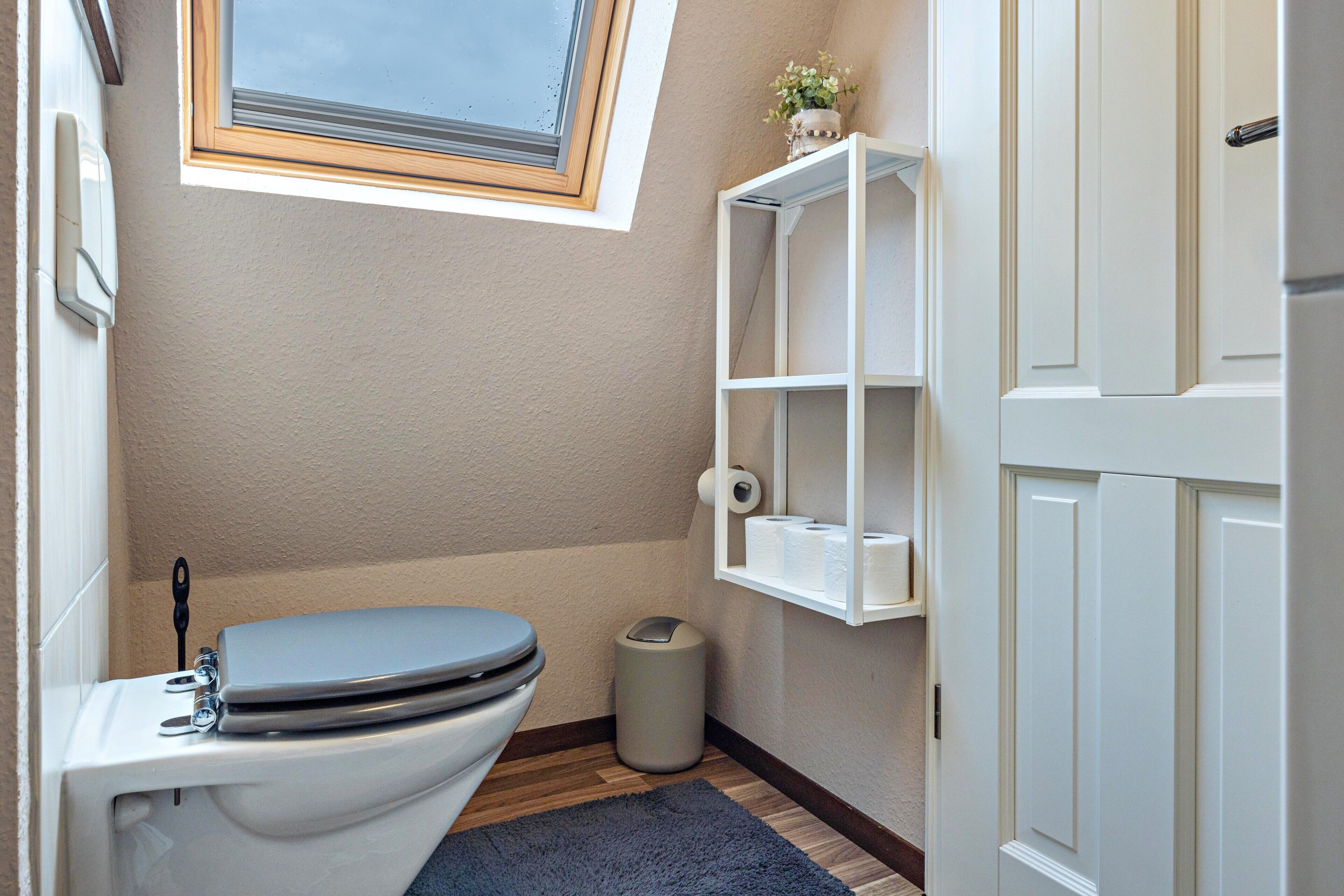 A bathroom with a toilet, a trash can, a shelf with toiletries, and a window above the toilet.