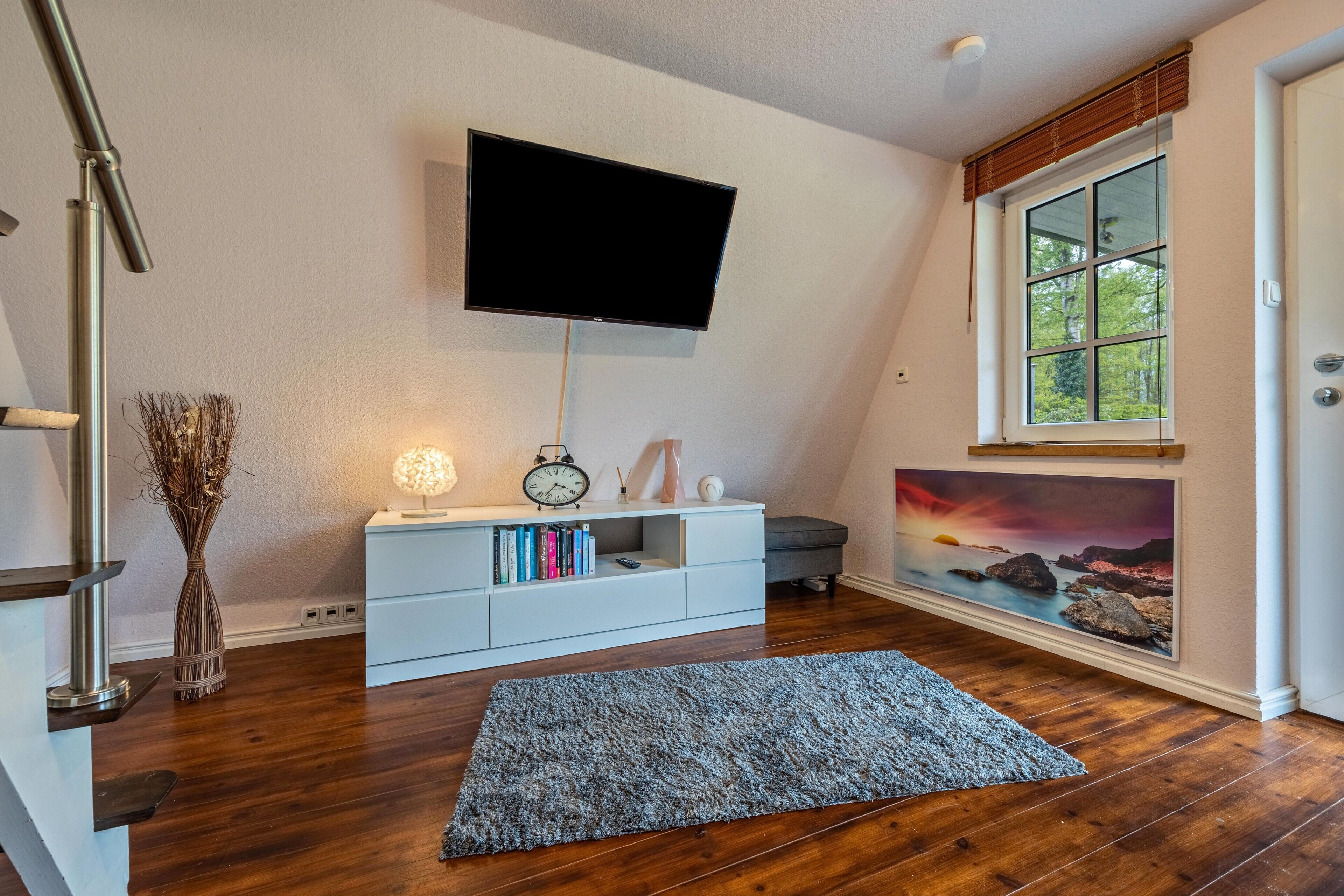 A modern living room with a flat-screen TV mounted on the wall, a white console table with books, a decorative vase, and a painting of a sunset.