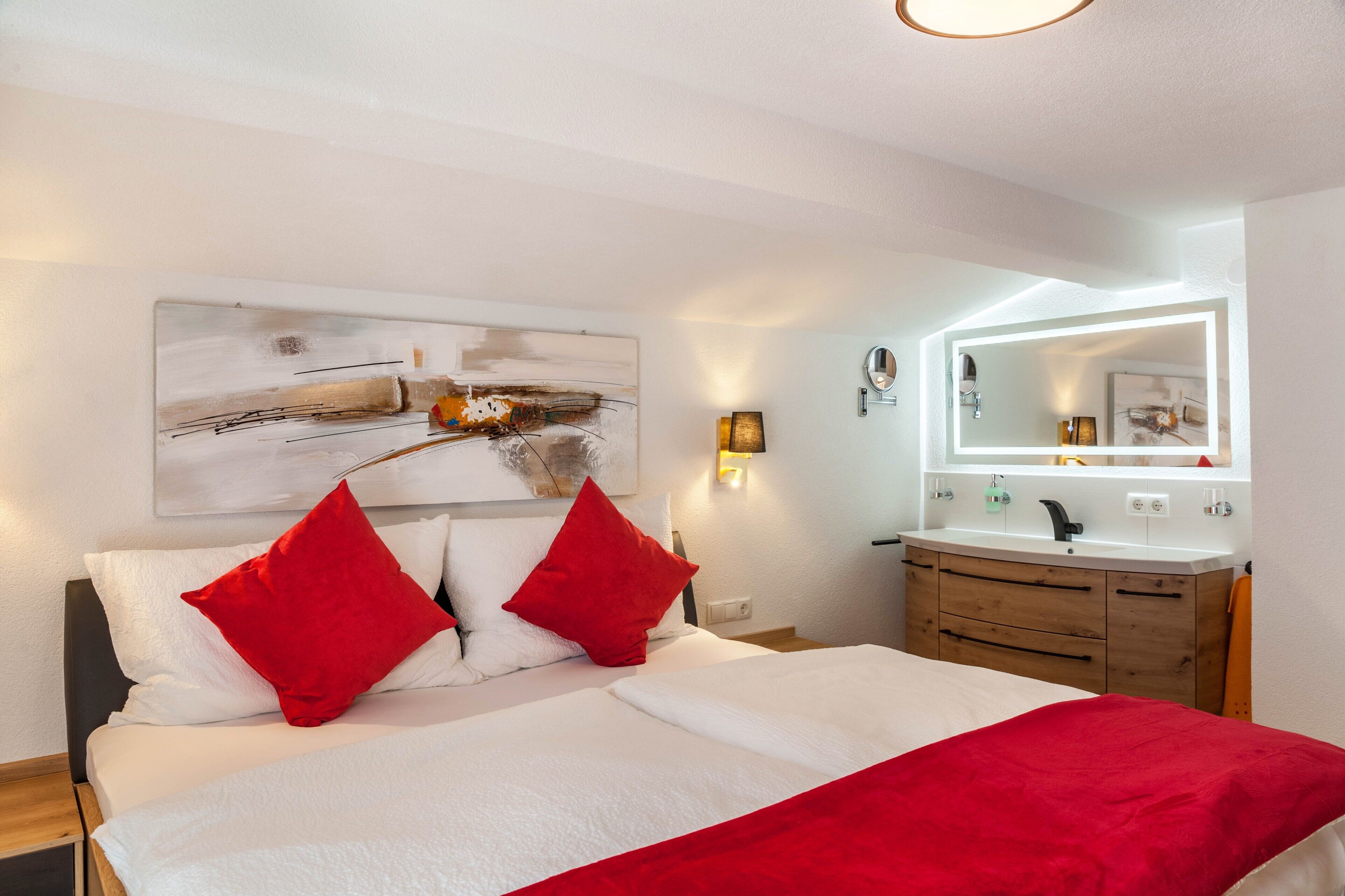A bedroom with a bed, red pillows, a bedside table, a mirror, and a wall-mounted artwork.