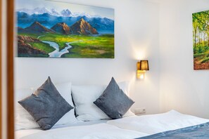 Hotellihuone, jossa on sänky, kaksi tyynyä, vuoristomaisemaa esittävä taulu ja seinään kiinnitetty valaisin.