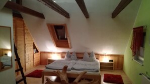Un dormitori amb sostre inclinat, bigues de fusta, un llit amb roba de llit blanca, dues taules de nit, una catifa vermella i un capçal de llit de fusta.