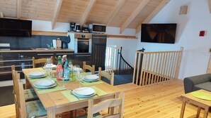 Un coin repas avec une table en bois dressée pour quatre personnes, une cuisine équipée d’appareils électroménagers modernes et un téléviseur fixé au mur.