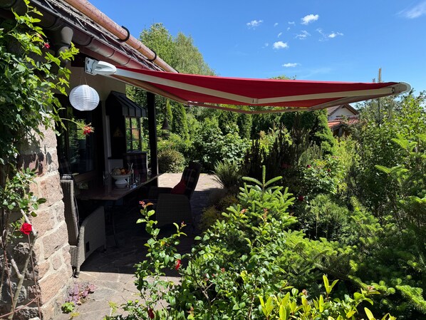 Un jardín con un muro de piedra, un toldo rojo y blanco, y abundante vegetación.