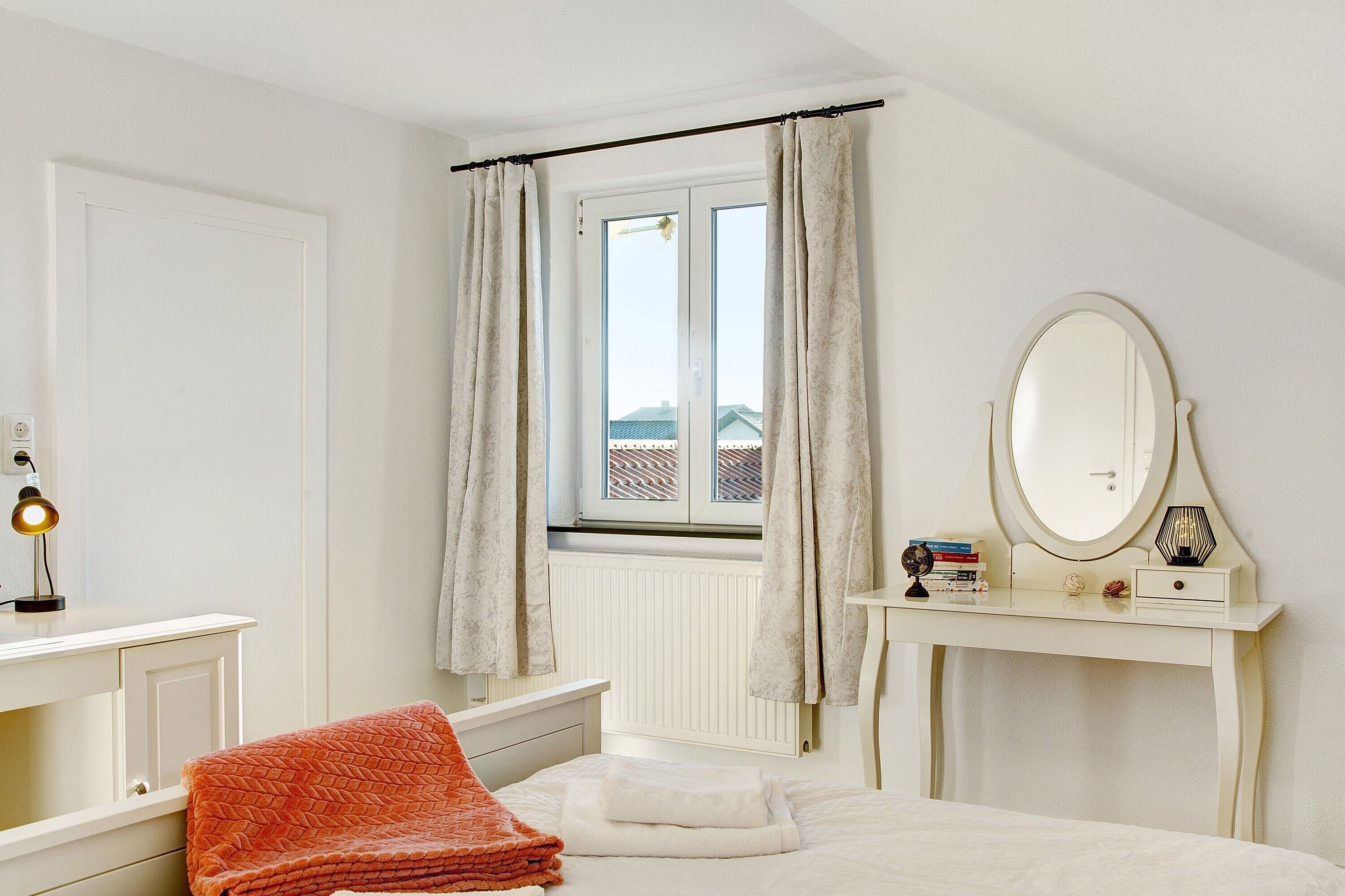 Ein Schlafzimmer mit Bett, Nachttisch, Spiegel, Fenster mit Vorhängen und Heizkörper.