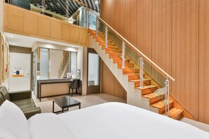 Ein modernes Hotelzimmer mit einem großen Bett, einem Schreibtisch, einem Sessel und einer Treppe mit Holzhinweisstufen und Glasgeländern.