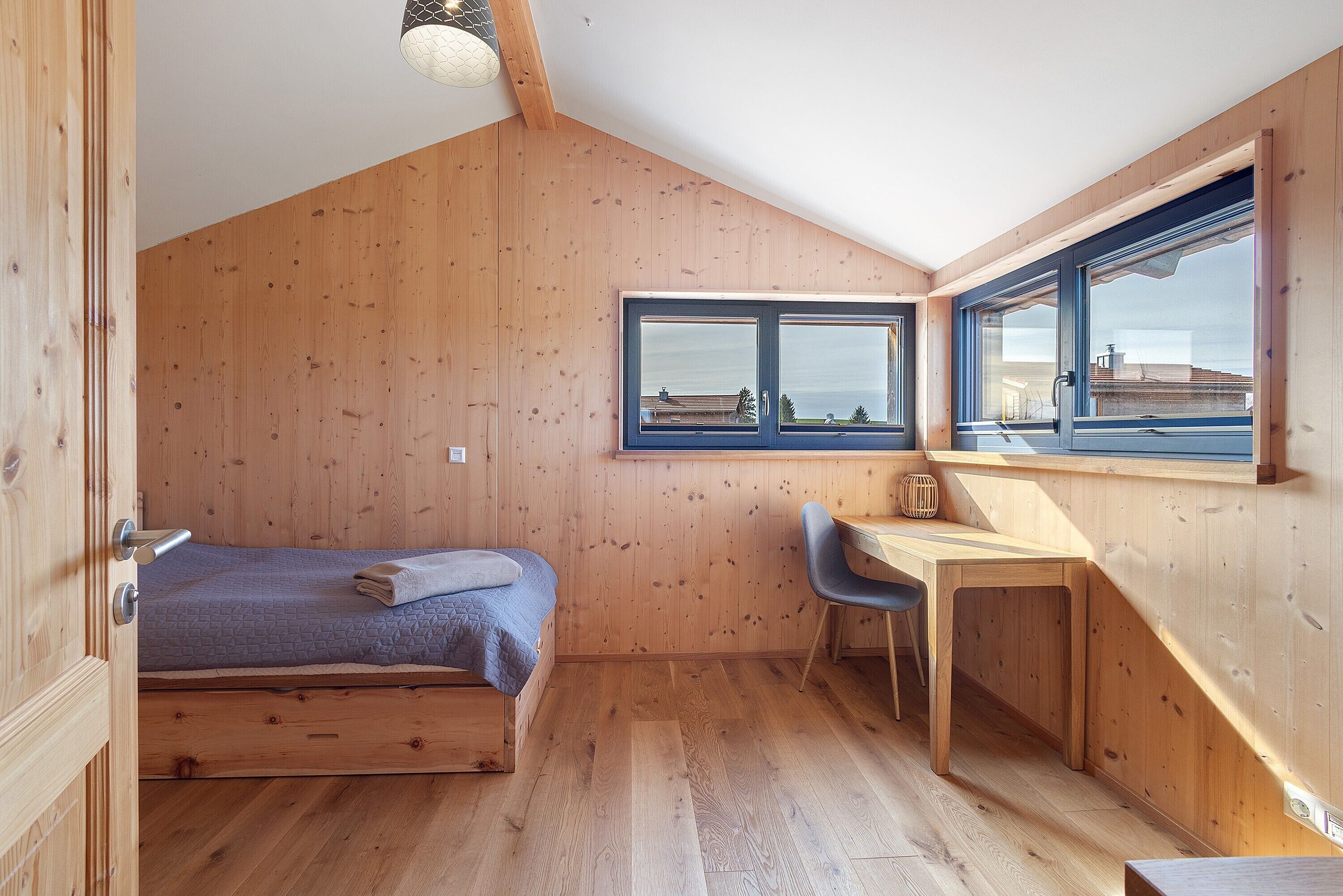 A small, wooden room with a bed, a desk, and a chair. There are windows providing natural light.