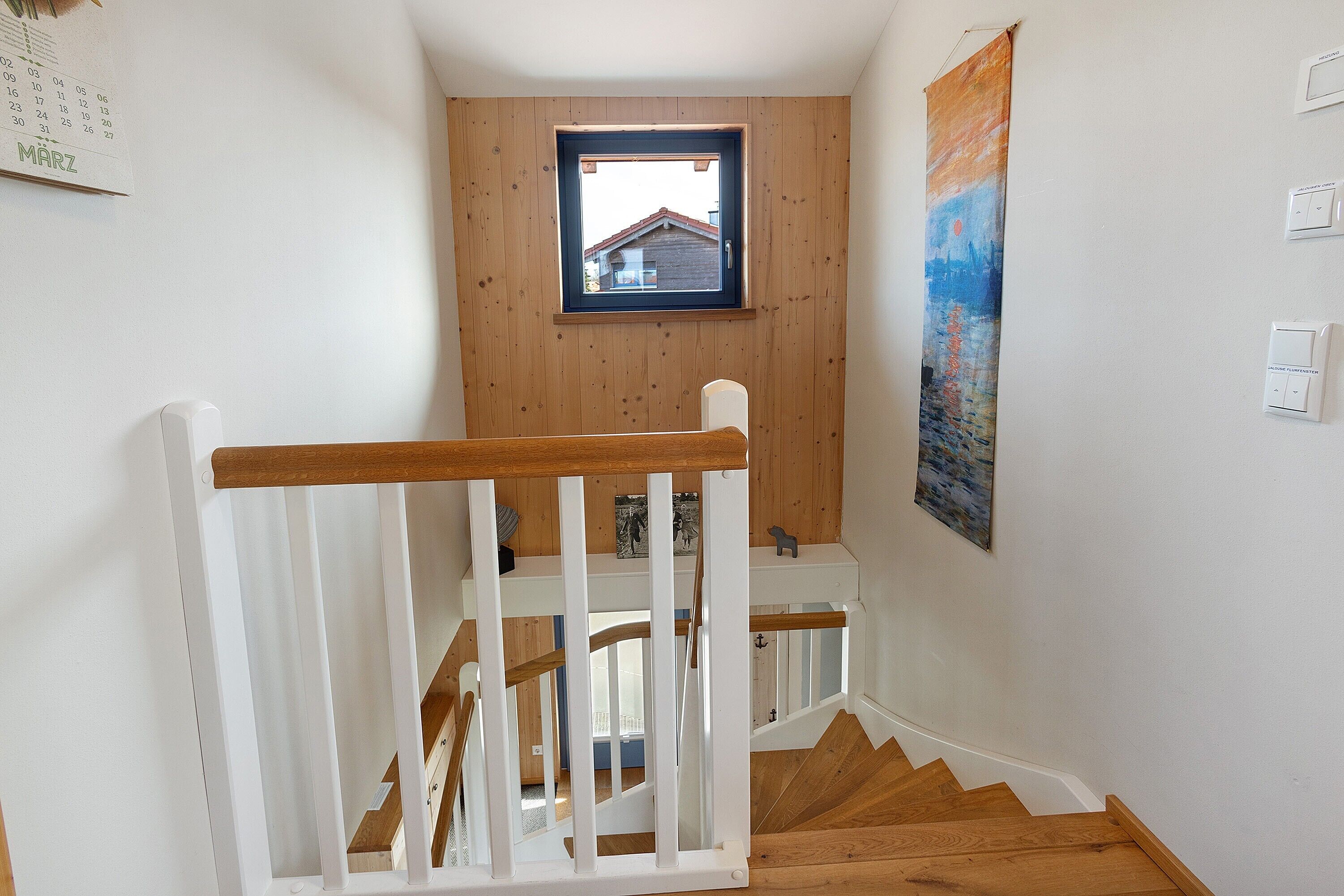 En trätrappa med ett vitt räcke, ett litet fönster med utsikt över ett hus och en kalender på väggen.