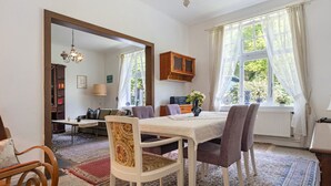 A dining area with a table, chairs, a chandelier, a window with curtains, and a bookshelf in the background.