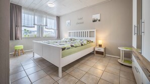 Ein Schlafzimmer mit einem Bett, Nachttischen, einem Fenster mit Vorhängen, einem kleinen Hocker und einem wandmontierten Spiegel.