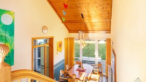 Ein Essbereich mit einem Holztisch und -stühlen, einer Decke mit hängenden Lampen und Blick auf einen Garten durch große Fenster.