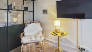 A room with a wicker chair, a small round table with a lamp, a black TV on the wall, and a black shelving unit with a mirror. - "Marisol" - 700m from the sea by Interhome (Skagen)
