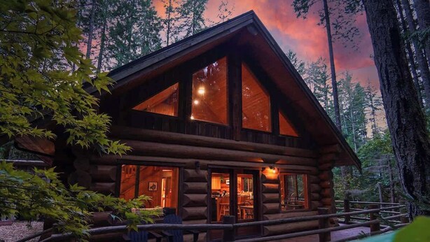 Une cabane en rondins dotée de grandes fenêtres, d’un porche et donnant sur une forêt.