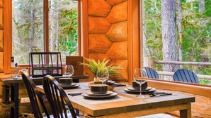 L’intérieur d’un chalet en bois, avec une table à manger dressée pour deux personnes, de grandes fenêtres et une vue sur une forêt.