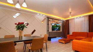 A modern living room with a dining area, orange sofas, a TV mounted on the wall, and a vase with red flowers on the table. - Lux Suites Nakuru VIP Suites (Nakuru)