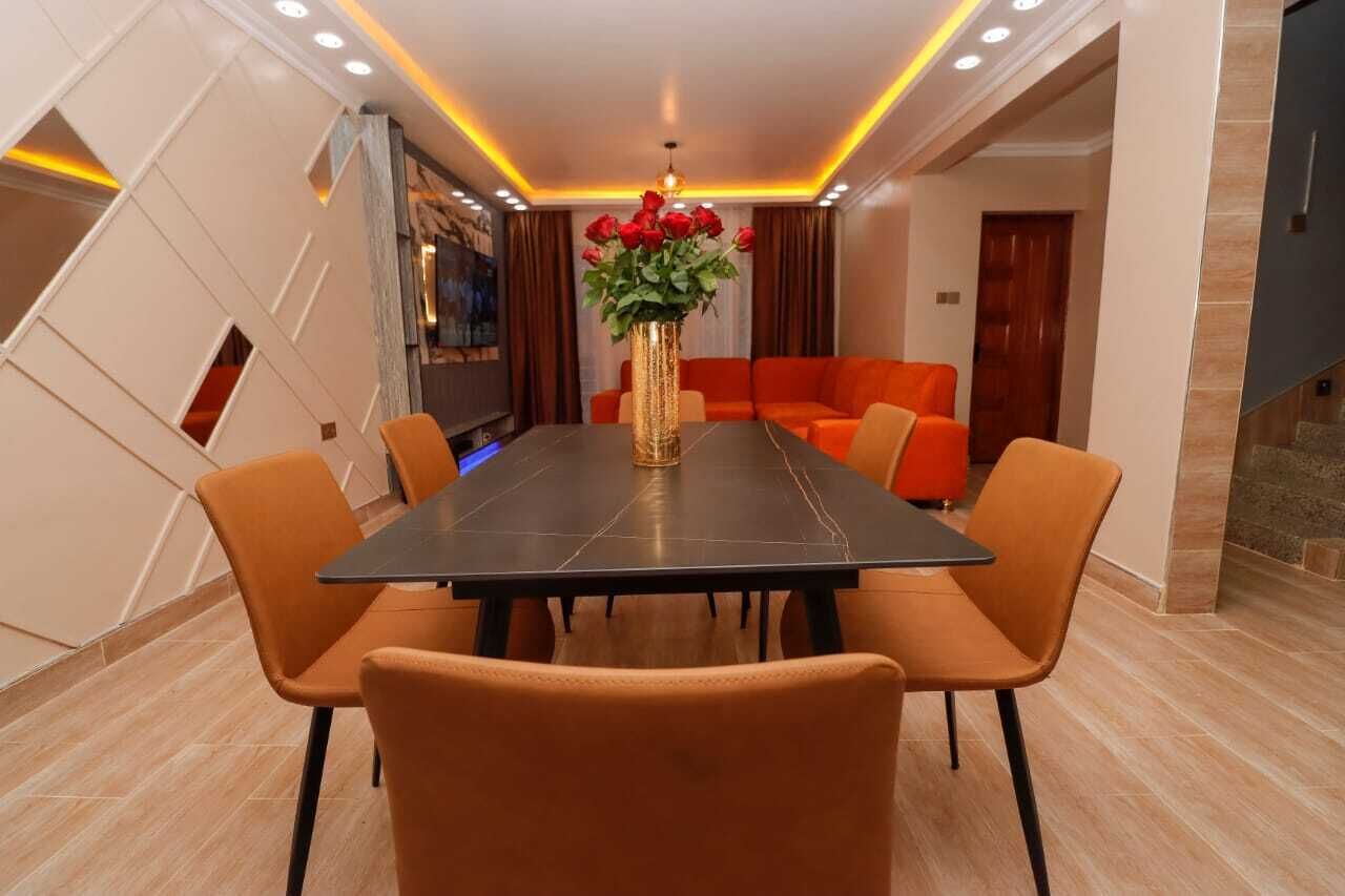 Ein Essbereich mit einem rechteckigen Tisch, sechs orangefarbenen Stühlen, einer Vase mit roten Blumen und einer modernen Decke mit Einbuchtungslampen.