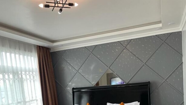 A hotel room with a large bed, two pillows, a bedside table, a nightstand, a window with curtains, and a ceiling light fixture. - Lux Suites Nakuru VIP Suites (Nakuru)