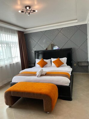 A hotel room with a large bed, two pillows, a bedside table, a nightstand, a window with curtains, and a ceiling light fixture. - Lux Suites Nakuru VIP Suites (Nakuru)