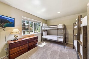 A room with a bunk bed, a desk with a lamp, a window with a view of a forest and mountains, and a painting on the wall.