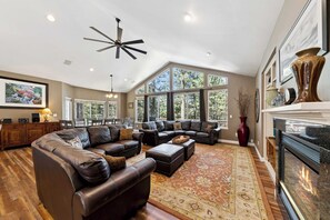 A spacious living room with a large L-shaped sectional sofa, a fireplace, and a dining area with a view of the outdoors. - HUGE 7Br (+Bonus Room) Cabin, Pool Table, Games (South Lake Tahoe)