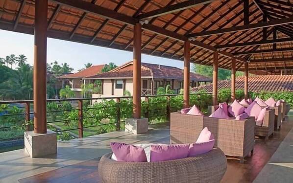 A covered outdoor seating area with wicker furniture and purple cushions, overlooking a residential area with houses and greenery.