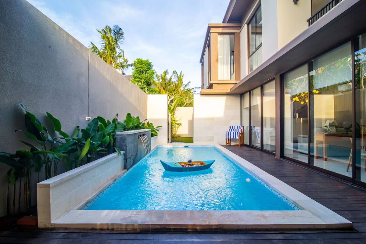 A modern swimming pool with a boat, surrounded by lush greenery and a contemporary building.