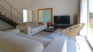 A modern living room with a sofa, a coffee table, a TV, and a wicker chair. There is a staircase on the left and a large window on the right. - Vacation home Caleu by Interhome (Binissalem)