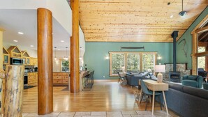 A spacious living area with a wooden ceiling, a stone fireplace, and large windows offering a view of the outdoors. - 4000sqft, 1-acre Custom Cabin Retreat w Game Room, Hot Tub & Sauna @Mt Hood (Rhododendron)