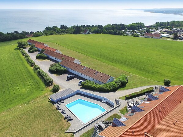 An aerial view of a property with a swimming pool, a long building with a red roof, and a large green field.