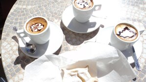 Una mesa con tres tazas de café, cada una con un diseño diferente en la espuma, y un pastel sobre una servilleta.