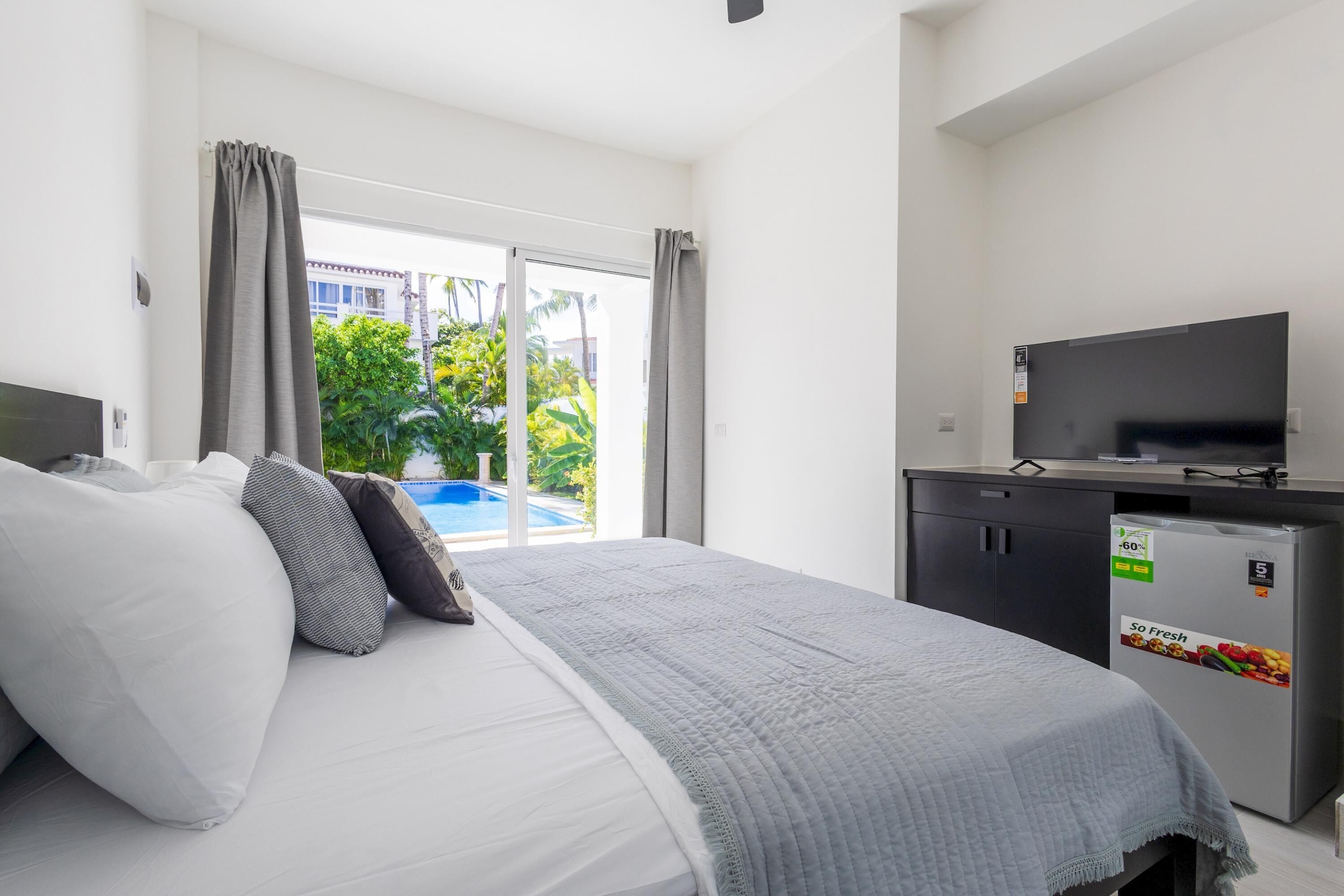 A hotel room with a bed, a TV, a refrigerator, and a view of a swimming pool.