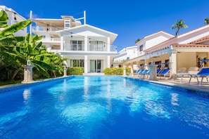 A swimming pool with a clear blue water, surrounded by white buildings, palm trees, and lounge chairs.