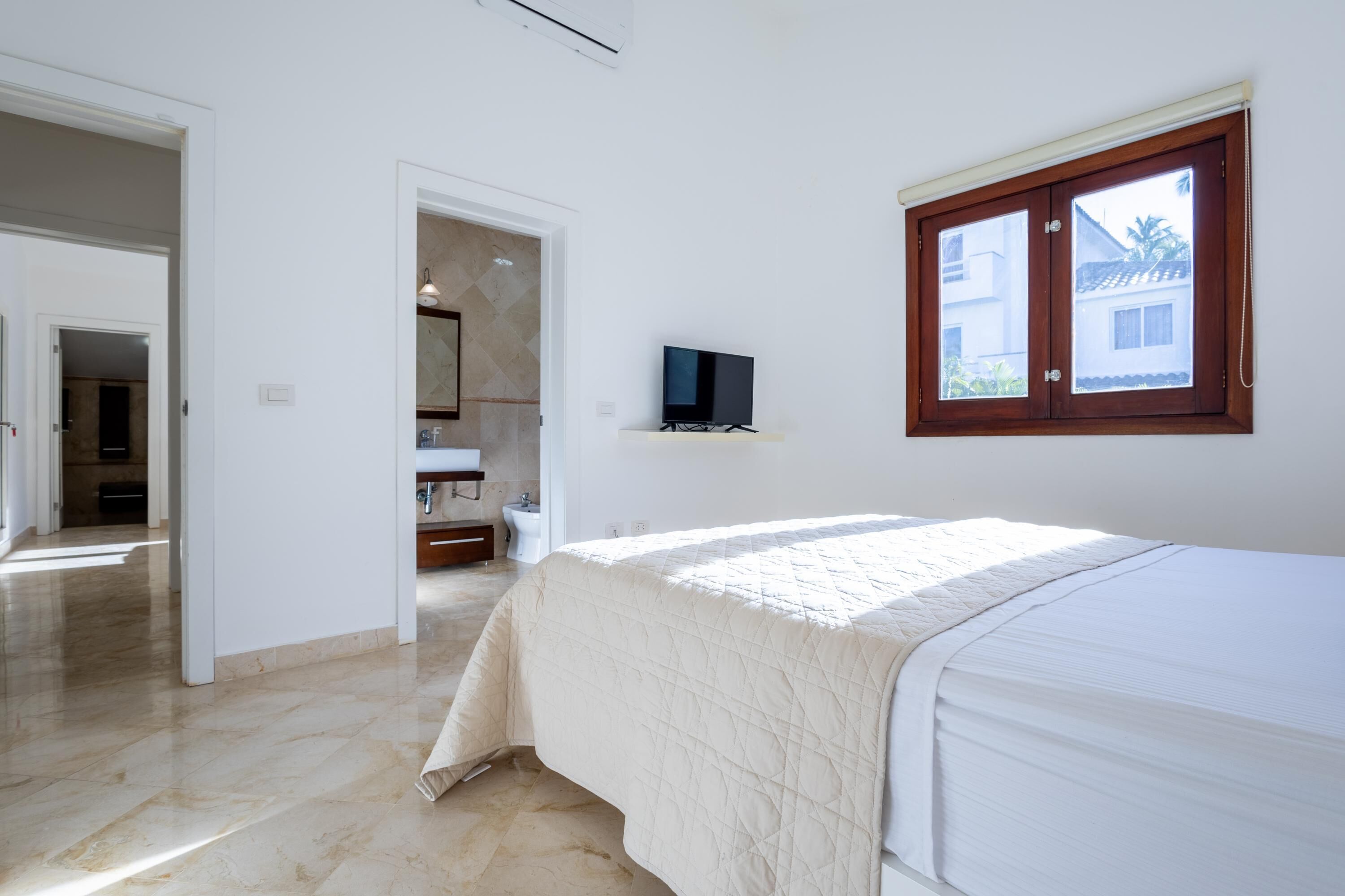 A bedroom with a bed, a television on a shelf, and a window with a view of a building outside.