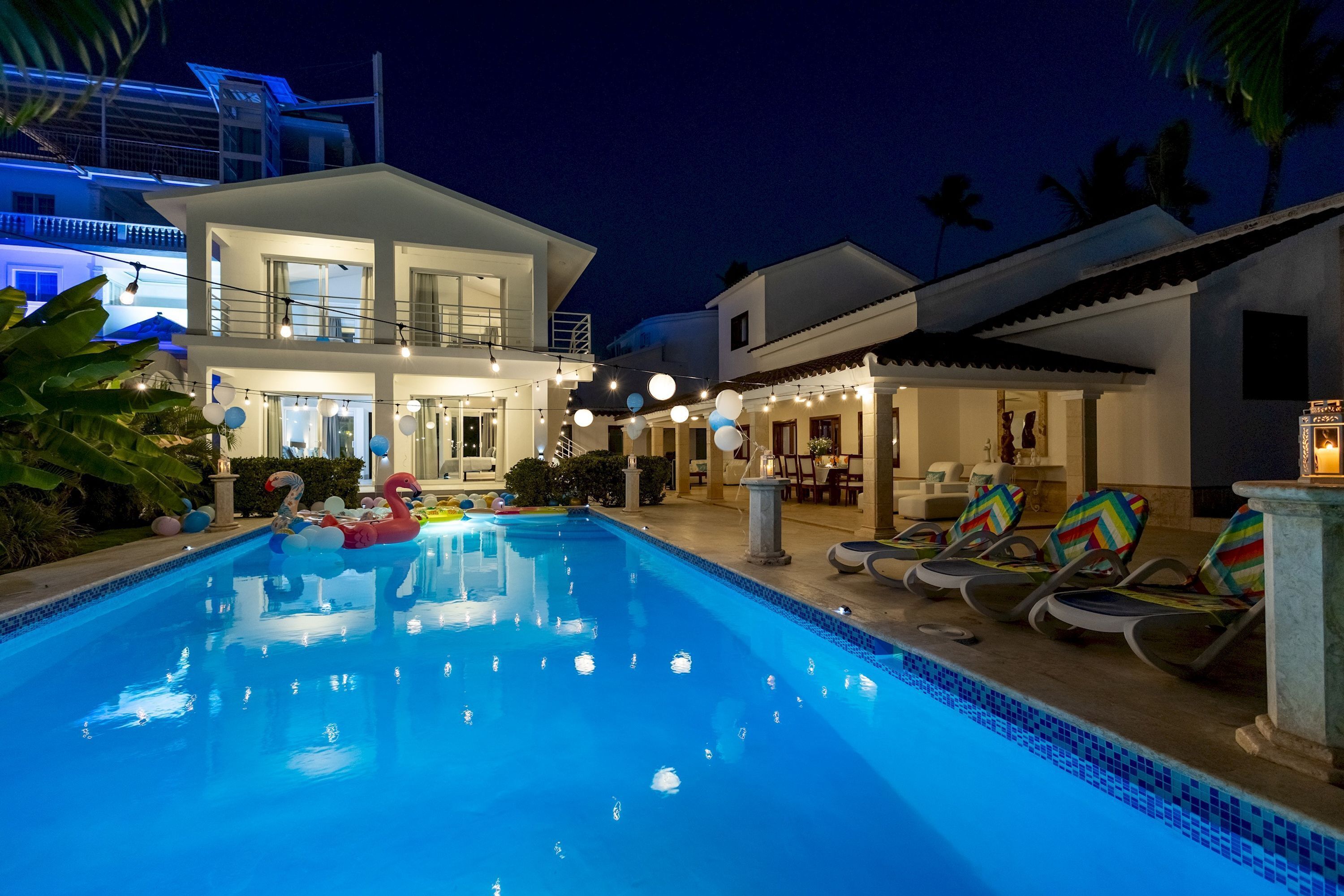 A swimming pool with lounge chairs, a house with balconies, and string lights.