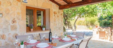 An outdoor dining area with a stone wall, wooden beams, and a view of greenery.