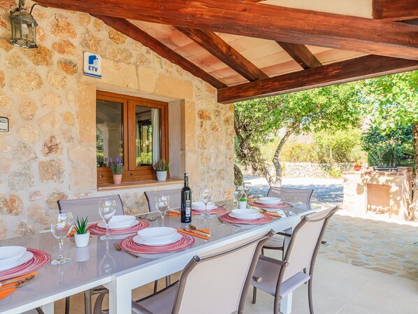 An outdoor dining area with a stone wall, wooden beams, and a view of greenery.