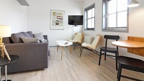 Un salon moderne avec un canapé gris, une petite table blanche et deux chaises. Un téléviseur est fixé au mur, il y a une fenêtre avec des stores et un luminaire suspendu.
