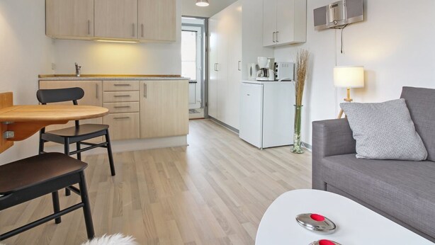 Une cuisine moderne avec des meubles en bois, un coin repas avec une table et des chaises, un canapé gris et une petite table blanche agrémentée d’objets rouges.