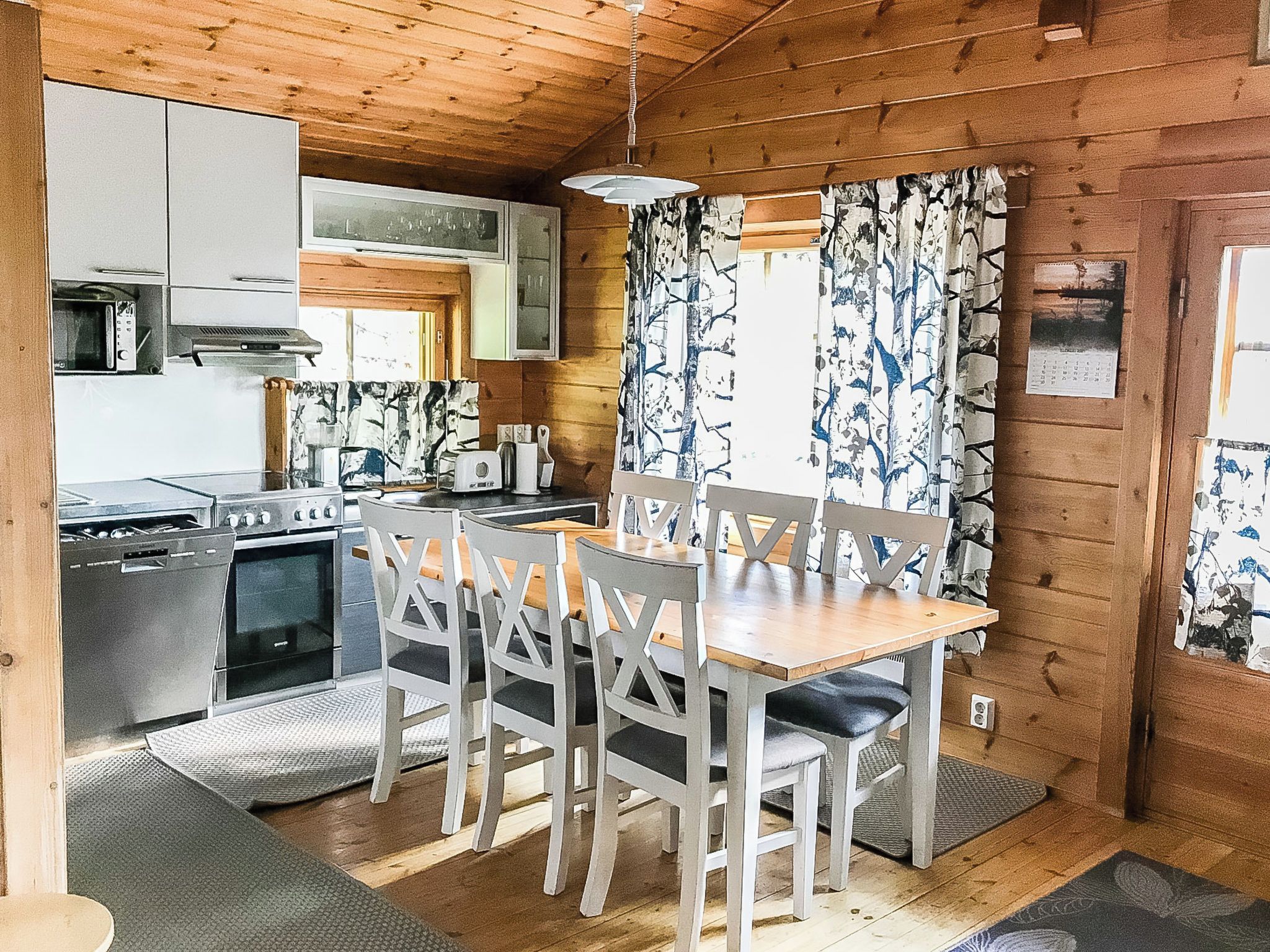 A compact kitchen with a dining area, wooden walls, and a window with patterned curtains.