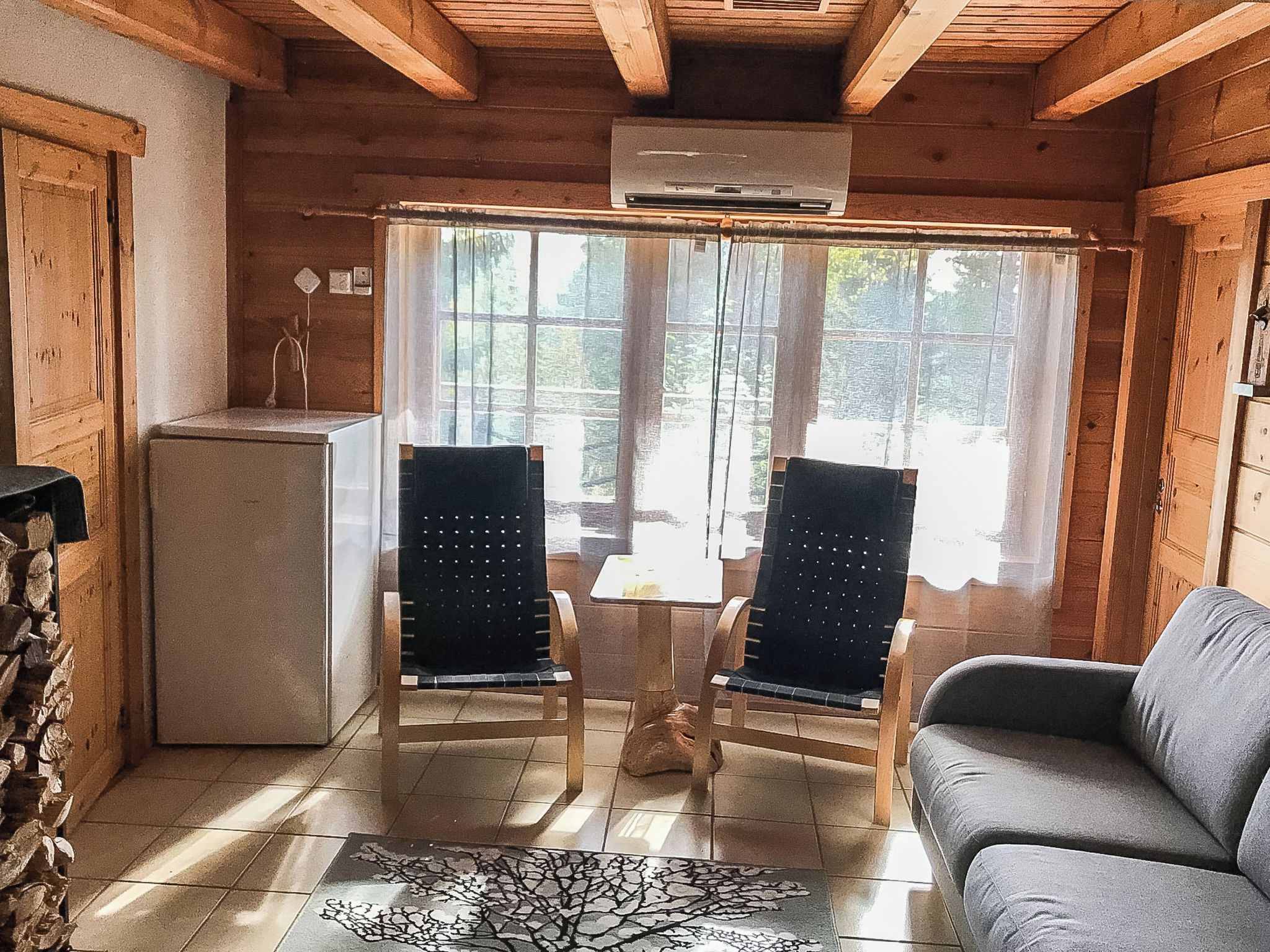 A log cabin interior with a stone wall, a gray sofa, two chairs, a small table, and a window with a view of trees.